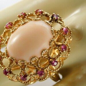 May include: A gold ring featuring a large, oval, peach-colored gemstone at the center, surrounded by a decorative gold filigree and small, round, red gemstones. The ring is set against a neutral background.