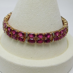 May include: A gold-toned bracelet featuring two parallel rows of oval-cut, deep pink gemstones. The links are connected by small, silver-toned accents embedded with tiny, clear, sparkling stones, creating a continuous, symmetrical design.
