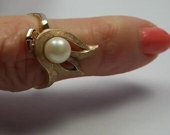 May include: A gold-toned ring featuring a central, round pearl. The ring's design includes stylized, leaf-like elements surrounding the pearl. The ring is worn on a finger with a coral-colored nail polish.