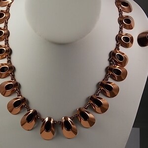 May include: A copper-toned necklace and matching earrings set. The necklace features a series of open, teardrop-shaped links connected by small chains. The earrings have a similar design. The jewelry is displayed on a white mannequin bust.