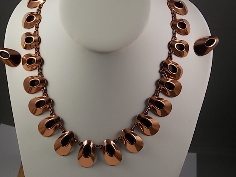 May include: A copper-toned necklace and matching earrings set. The necklace features a series of open, teardrop-shaped links connected by small chains. The earrings have a similar design. The jewelry is displayed on a white mannequin bust.