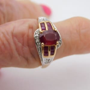 May include: A two-tone ring with a large oval ruby gemstone as the focal point. The ring features smaller rectangular rubies and clear accent stones. The band is silver and gold in color.