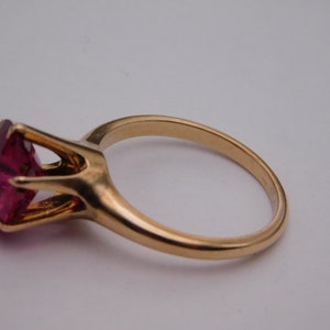 May include: A gold-colored ring featuring a large, faceted, magenta-colored gemstone. The gemstone is held in place by a four-prong setting. The ring band is smooth and rounded.