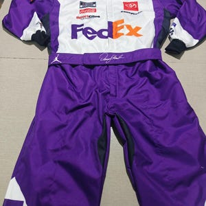 May include: A purple and white racing suit featuring the FedEx logo on the chest and collar. The suit includes various sponsor logos such as NASCAR Cup Series, Coca-Cola, SportClips, and Goodyear, along with Jordan brand logos and a signature on the belt.