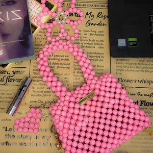 Puede incluir: Un bolso rosa con forma de coraz&oacute;n hecho de cuentas, con un cierre dorado y un llavero de flores. Tambi&eacute;n se ven un libro y un l&aacute;piz labial.