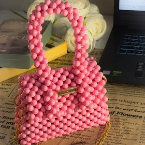 Puede incluir: Bolso de mano rosa con cuentas y correa dorada. El bolso est&aacute; hecho de peque&ntilde;as cuentas redondas y tiene forma rectangular. El bolso est&aacute; sobre un peri&oacute;dico. El dise&ntilde;o del bolso es llamativo.
