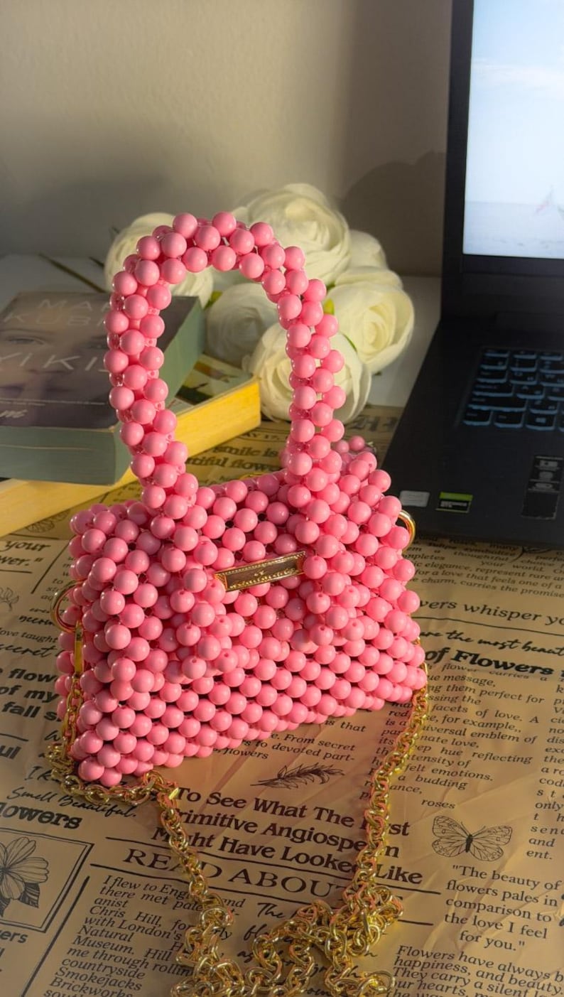 Puede incluir: Bolso de mano rosa con cuentas y correa dorada. El bolso est&aacute; hecho de peque&ntilde;as cuentas redondas y tiene forma rectangular. El bolso est&aacute; sobre un peri&oacute;dico. El dise&ntilde;o del bolso es llamativo.