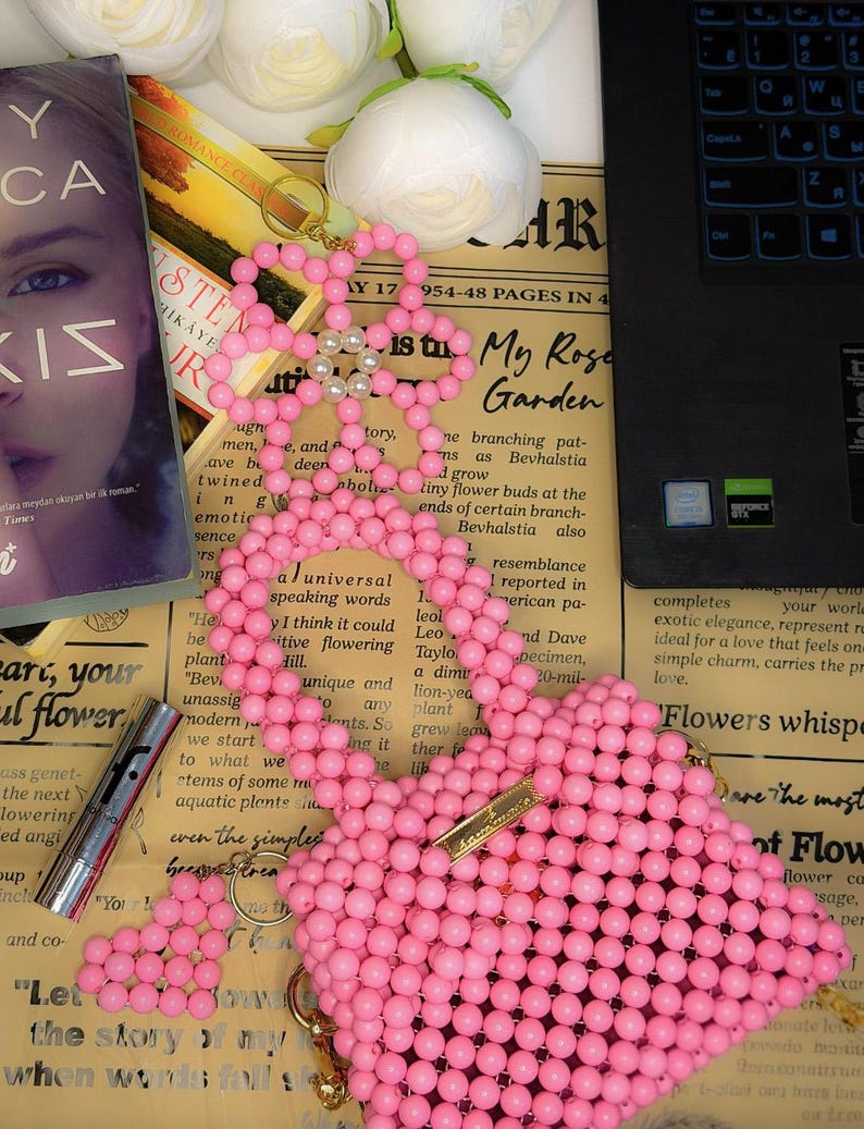 Puede incluir: Un bolso rosa con forma de coraz&oacute;n hecho de cuentas, con un cierre dorado y un llavero de flores. Tambi&eacute;n se ven un libro y un l&aacute;piz labial.