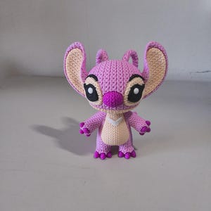 May include: A handmade crochet figure of a character with large ears, large black eyes, and a purple body. The figure has a light pink face and ears, with a darker purple nose and accents. The figure is standing on a light gray surface.