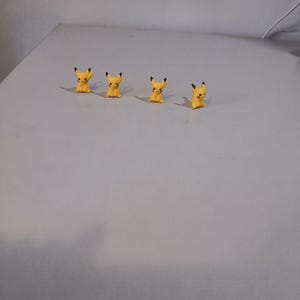 Op de afbeelding: Vier kleine, gele Pikachu-beeldjes met zwartgepunte oren en rode wangen zijn in een rij gerangschikt op een lichtgrijze ondergrond. De beeldjes zitten en kijken naar voren. De achtergrond is effen lichtgrijs.