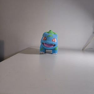 Op de afbeelding: Een gebreide Bulbasaur-figuur in blauw, groen en roze. De figuur heeft rode ogen, een lachende mond en een groene bol op zijn hoofd. Het staat op een wit oppervlak.