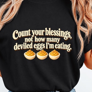 May include: A black short-sleeve t-shirt featuring the text "Count your blessings, not how many deviled eggs I'm eating." in a cream-colored font, accompanied by an illustration of three deviled eggs with yellow yolks.