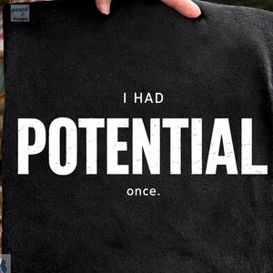 Pode incluir: Um tecido quadrado preto com as palavras "I HAD POTENTIAL once." em letras brancas grandes. O tecido é segurado por uma mão, com um fundo desfocado ao ar livre visível.