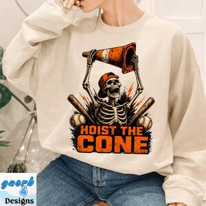 May include: Beige sweatshirt with a graphic of a skeleton wearing a baseball cap and holding a traffic cone. The text "HOIST THE CONE" is below the skeleton, with baseballs and bats on either side.