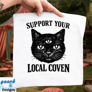 Peut inclure: Coussin carré blanc avec une illustration noire d'un chat à trois yeux. Le texte "SUPPORT YOUR LOCAL COVEN" est imprimé au-dessus et en dessous de l'image du chat en gras noir. Le coussin est tenu par une main.