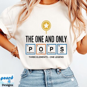 Puede incluir: Una camiseta color crema con un gráfico que incluye un icono de estrella dorada y el texto THE ONE AND ONLY. Debajo, cuatro cuadrados estilo tabla periódica deletrean POPS con los elementos Fósforo, Oxígeno, Fósforo y Azufre. Texto: THREE ELEMENTS • ONE LEGEND.