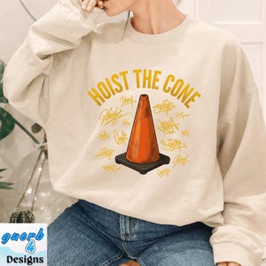 May include: Cream-colored sweatshirt with the phrase "HOIST THE CONE" in gold lettering above an orange traffic cone graphic. The cone is on a black base, surrounded by various signatures in gold.