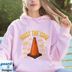 May include: A light pink hoodie with the words "HOIST THE CONE" in gold lettering and a graphic of an orange traffic cone. The cone is surrounded by various signatures. The hoodie has a drawstring hood and a front pocket.