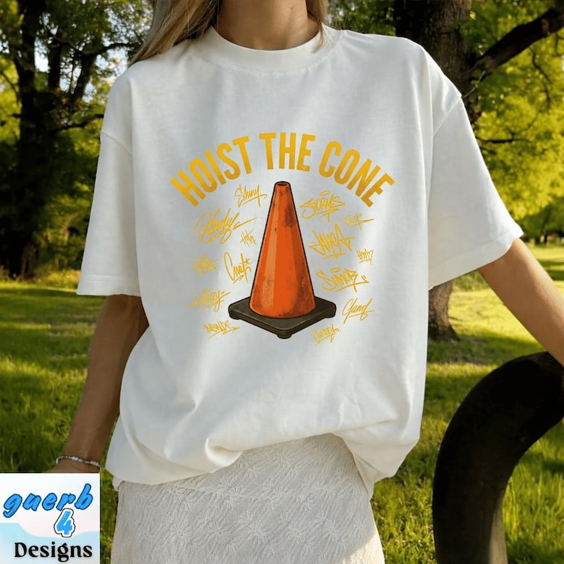 May include: White t-shirt with the text "HOIST THE CONE" in yellow, featuring an orange traffic cone graphic. The design includes various yellow signatures around the cone. The shirt is a casual, unisex style.