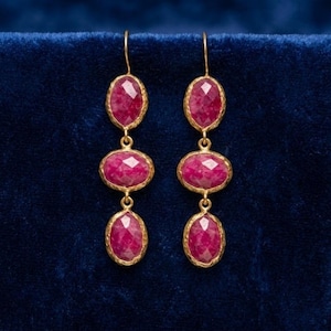 May include: A pair of gold-colored earrings with three oval, faceted, ruby-colored gemstones. The earrings are displayed against a dark blue background. The gemstones are set in a gold-colored bezel setting.