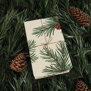 May include: A wrapped gift with cream-coloured paper featuring a green pine branch and pine cone pattern. The gift is tied with twine and rests on a bed of green pine needles and several brown pine cones. A festive, nature-themed presentation.