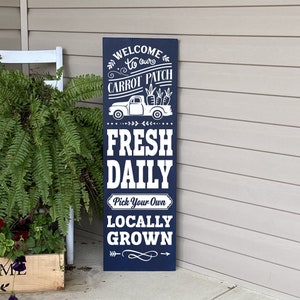 May include: A blue wooden sign with white text that reads "Welcome to our Carrot Patch, Fresh Daily, Pick Your Own, Locally Grown". The sign features a white silhouette of a pickup truck carrying carrots.