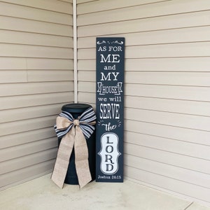 May include: Black wooden sign with white text that reads "As for me and my house we will serve the Lord Joshua 24:15". The sign is leaning against a beige wall.