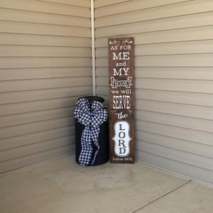 May include: A wooden sign with the text "As for me and my house we will serve the Lord Joshua 24:15" in white lettering. The sign is brown and has a distressed look. It is leaning against a wall.