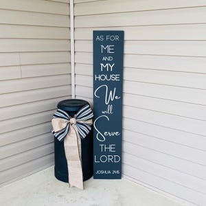 May include: A navy blue wooden sign with white lettering that reads "As for me and my house, we will serve the Lord, Joshua 24:15." A black metal container is decorated with a striped bow and a tan ribbon.