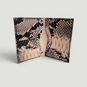 May include: A bi-fold card holder featuring a black and beige snakeskin pattern. The interior displays two vertical card slots with curved edges and a central pocket, all constructed with visible black stitching along the outer perimeter.