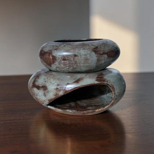 May include: A two-piece ceramic object with a smooth, rounded, pebble-like shape. The top section is a bowl-like piece, while the bottom section features a large, hollowed-out side opening. Both pieces have a mottled, teal-green and brown glaze.