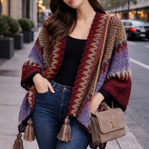 May include: A colorful, open-front knit poncho with a chevron pattern in shades of burgundy, purple, and tan. The poncho has long sleeves and is adorned with tassels. A brown handbag with a gold chain strap and a gold logo is also visible.