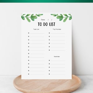 Puede incluir: Una lista de tareas imprimible en papel blanco con detalles de hojas verdes. La lista incluye secciones para una lista de tareas, las principales prioridades y recordatorios. La parte superior de la página dice "TO DO LIST" y "PRINTABLE A4 / A5 / LETTER". El papel descansa sobre una bandeja de madera.