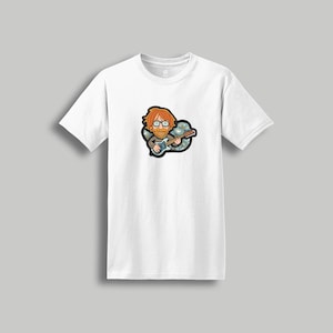 May include: White t-shirt featuring a cartoon illustration of a musician with red hair and glasses playing a guitar. The graphic is surrounded by a blue and green design. The shirt has short sleeves.