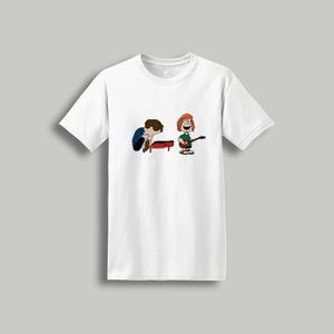 May include: White t-shirt featuring a cartoon illustration of two characters playing musical instruments. One character is playing a piano, and the other is playing a guitar. The characters are in a playful, cartoon style.
