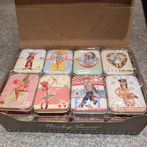 Puede incluir: Una caja de cartón llena de ocho latas de metal decorativas. Cada lata presenta una ilustración de estilo vintage con una variedad de temas, incluyendo ángeles, niños e imágenes patrióticas. Las latas son rectangulares con bordes redondeados.