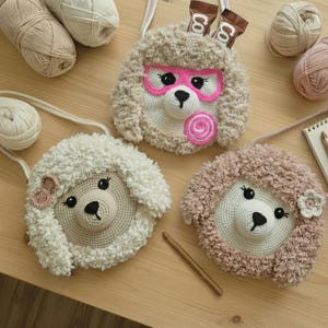 Op de afbeelding: Drie handgemaakte gehaakte tassen in de vorm van schapen, beige, wit en roze. Eén heeft een roze bril en een lolly, een andere een strik en de derde een bloem. De tassen zijn gemaakt van pluizig garen.