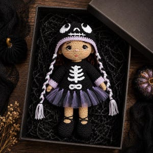 May include: A crocheted doll with tan skin, wearing a black hooded outfit featuring a white skeleton ribcage design and a purple tulle skirt. The doll has long braided hair with purple accents and is presented inside a black gift box.