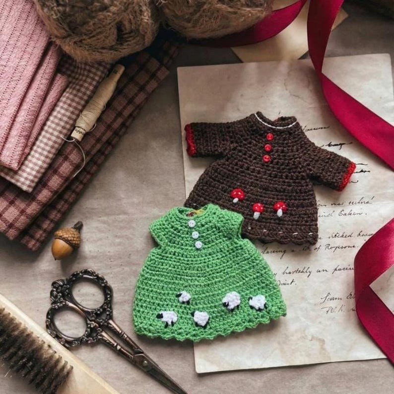 May include: Two miniature crochet dresses, one brown with red accents and mushroom details, the other green with white sheep. The dresses are on a vintage paper background with sewing supplies and fabric.