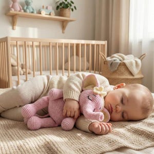 May include: A sleeping baby cuddles a pink crocheted elephant toy. The elephant has large ears, a trunk, and a yellow flower on its head. The baby is wearing a cream-colored sweater and is lying on a beige blanket.