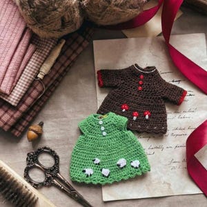 May include: Two miniature crochet dresses. One is brown with red trim and mushroom appliqu&eacute;s, the other is green with white sheep appliqu&eacute;s. The dresses are on a vintage paper background with other craft supplies.
