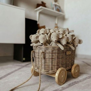 Puede incluir: Un vag&oacute;n de juguete de mimbre lleno de ratones de peluche de ganchillo beige. El vag&oacute;n tiene ruedas de madera y un asa de cuerda. Los juguetes son de color beige claro y tienen peque&ntilde;os ojos y narices marrones. El vag&oacute;n est&aacute; sobre una alfombra gris claro.
