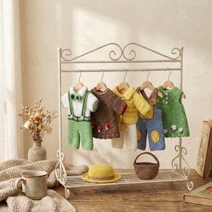 May include: A collection of handmade crochet baby clothes in various colors, including green, brown, and yellow, hanging on a decorative white rack. A yellow hat and a small brown basket sit on the rack's shelf. A mug and a blanket are in the foreground.