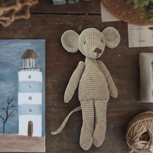 Puede incluir: Una mu&ntilde;eca de rat&oacute;n de ganchillo beige claro hecha a mano con orejas grandes, una cola larga y una nariz peque&ntilde;a de color marr&oacute;n oscuro. La mu&ntilde;eca est&aacute; de pie. Una pintura de un faro est&aacute; en el fondo.