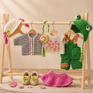 Peut inclure: Un portant en bois présente plusieurs tenues de bébé au crochet. Les articles comprennent un chapeau de lapin, un cardigan gris, un haut crème avec des fleurs roses, une salopette verte, un chapeau de grenouille, une jupe rose et une paire de chaussures.