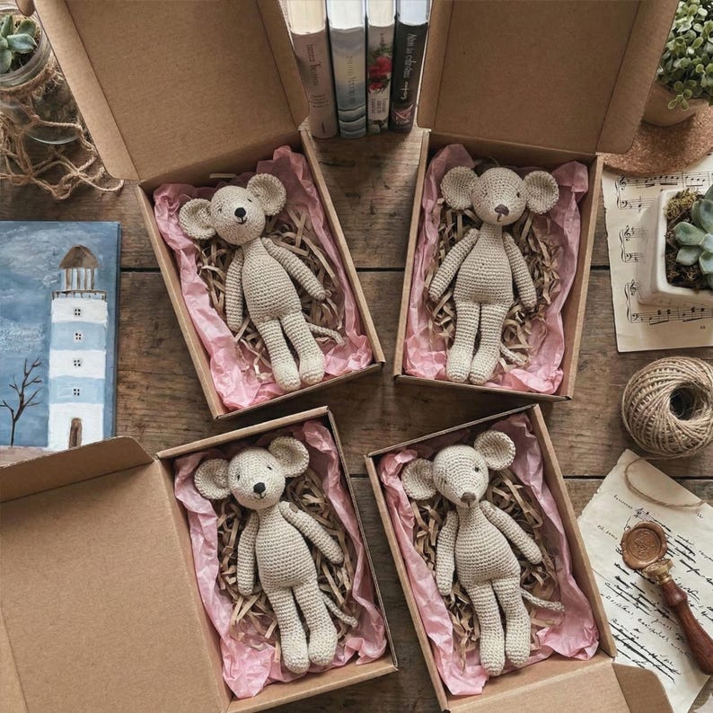 Puede incluir: Cuatro ratones de ganchillo beige, cada uno en una caja de cart&oacute;n forrada con papel rosa. Los ratones tienen orejas grandes y ojos negros peque&ntilde;os. Las cajas est&aacute;n dispuestas sobre una superficie de madera, con libros y objetos decorativos al fondo.
