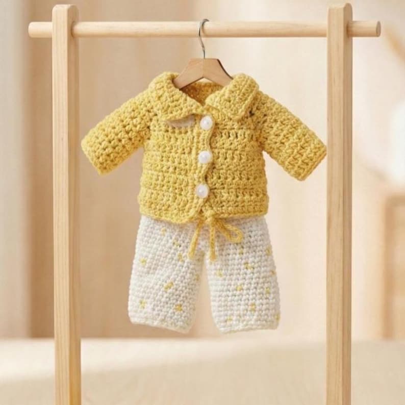 May include: A handmade crochet baby outfit in yellow and white. The set includes a button-up jacket and matching pants. The jacket has a collar and white buttons. The pants are white with yellow accents. The outfit is hanging on a wooden hanger.