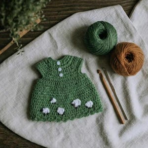 May include: A handmade green crochet baby dress with white sheep appliqu&eacute;s and three white buttons. Two crochet hooks and two balls of yarn, one green and one brown, are next to the dress. The dress is on a beige linen surface.