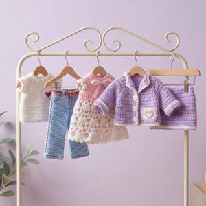 May include: A selection of handmade crochet doll clothes, including a white top, blue jeans, a pink dress, a purple jacket, and a skirt. Each item is displayed on a wooden hanger against a soft purple backdrop. The clothes feature heart-shaped details.