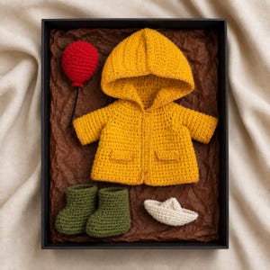 May include: A yellow crocheted hooded jacket, a pair of olive green crocheted booties, a small red crocheted balloon on a black string, and a small white crocheted paper-style boat, all arranged inside a black rectangular gift box.